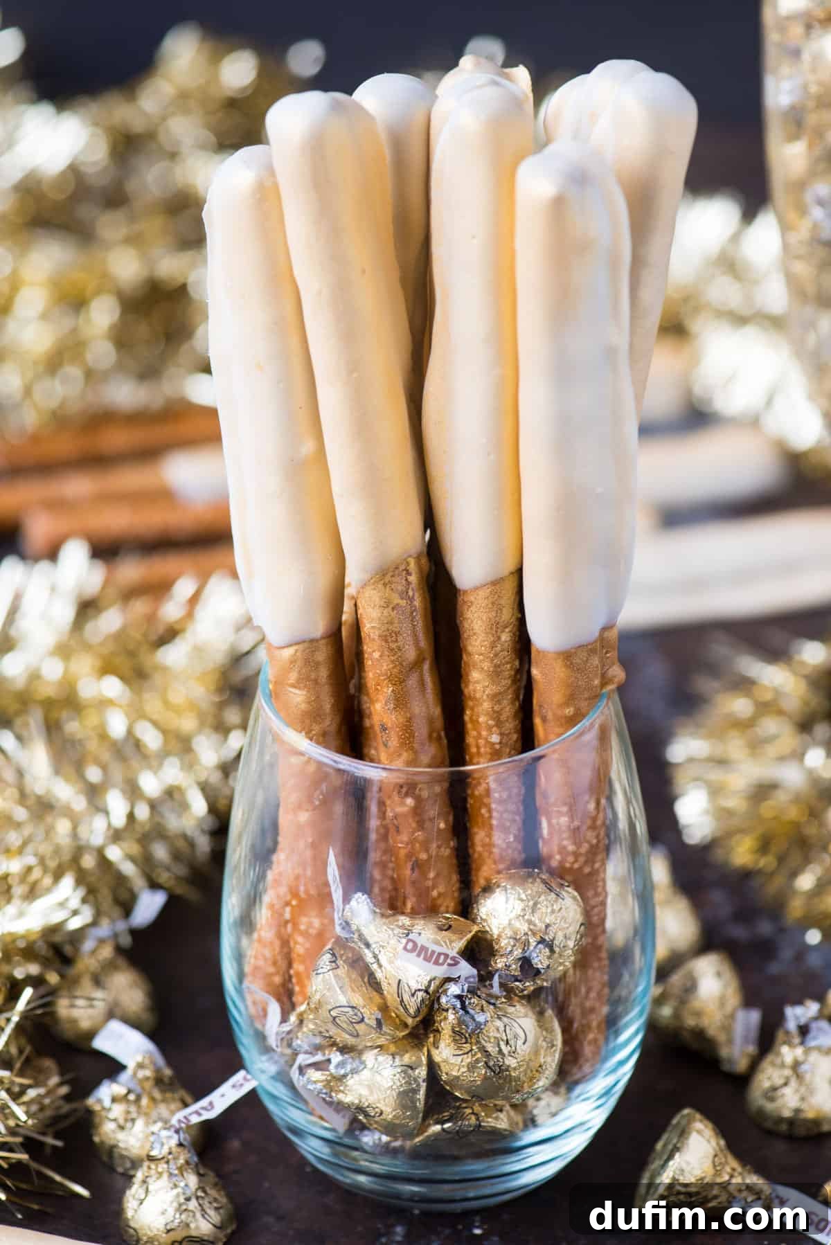 Elegant New Year's Eve gold pretzel rods, dipped in caramel and white chocolate, standing upright in a clear glass against a festive backdrop.