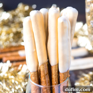 A close-up of New Years Eve pretzels—pretzel rods partially dipped in white chocolate, standing upright in a glass, with a blurred festive gold tinsel background.