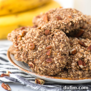 A plate of banana breakfast cookies with chopped nuts on top, stacked together. A striped cloth sits underneath the plate, and bananas are blurred in the background.