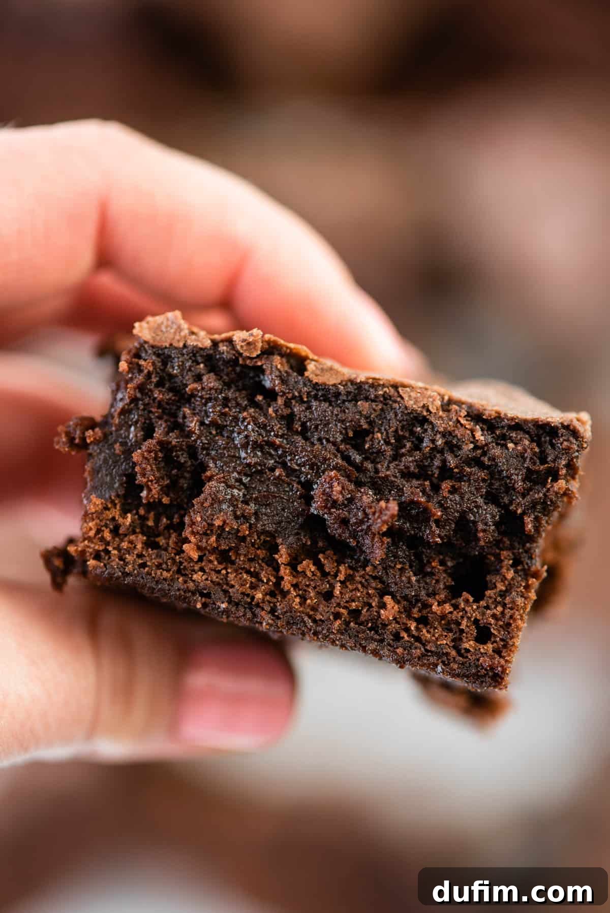 A hand holding a thick, rich slice of a fudgy brownie with a perfect crackled top, showcasing its moist texture.