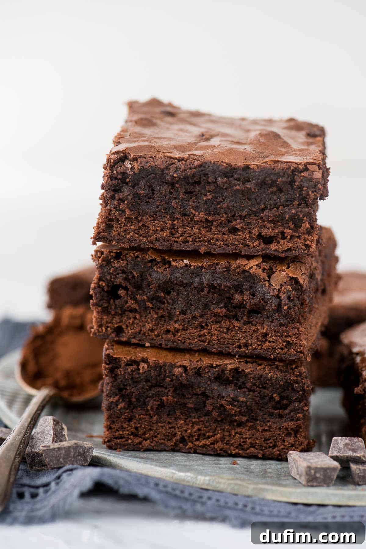 Three perfectly baked, rich chocolate brownies stacked on a gray plate, with a soft blue linen underneath, highlighting their decadent appearance.