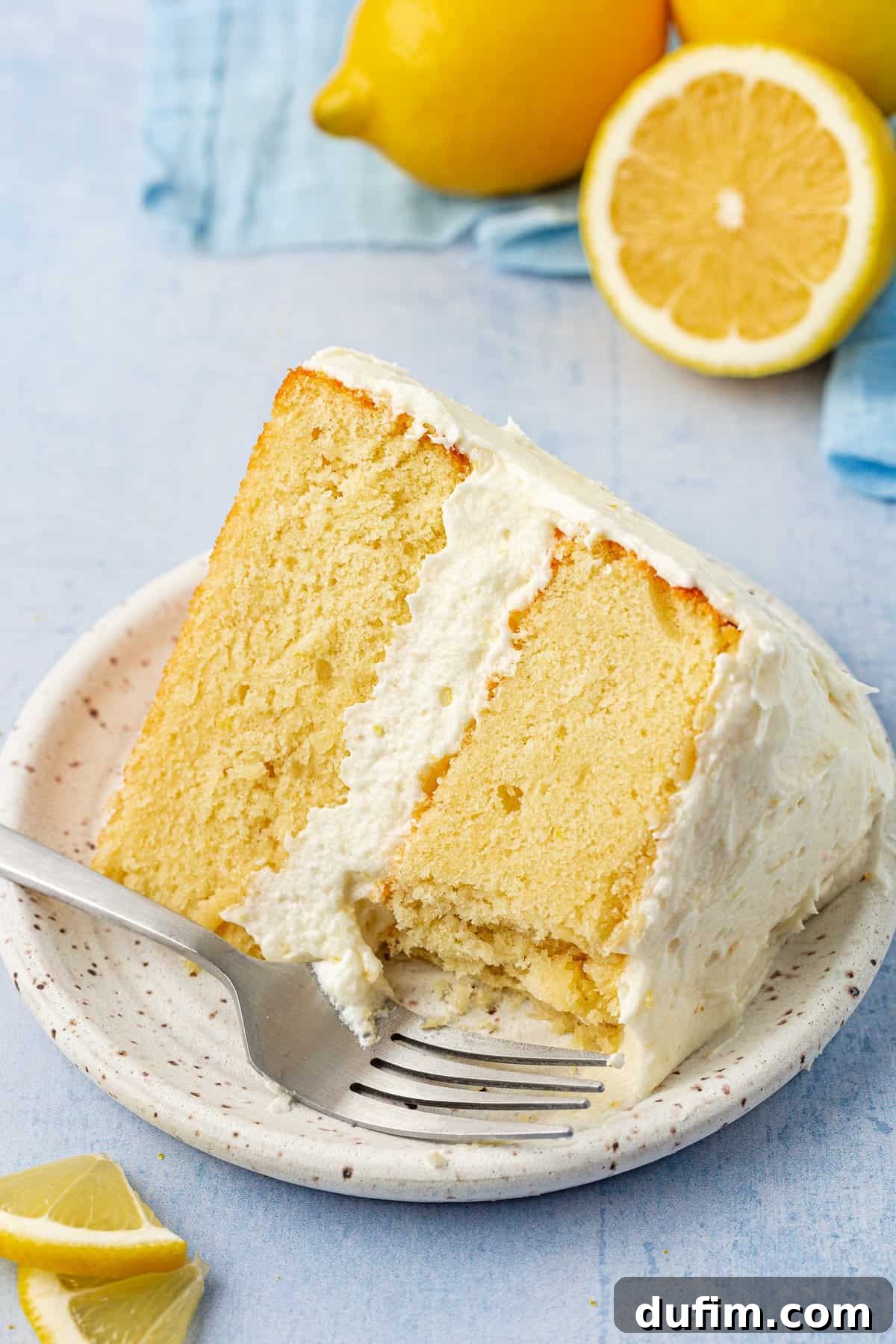 A delectable slice of lemon layer cake on a small white plate, accompanied by a fork and fresh lemon halves, showcasing the irresistible frosting.