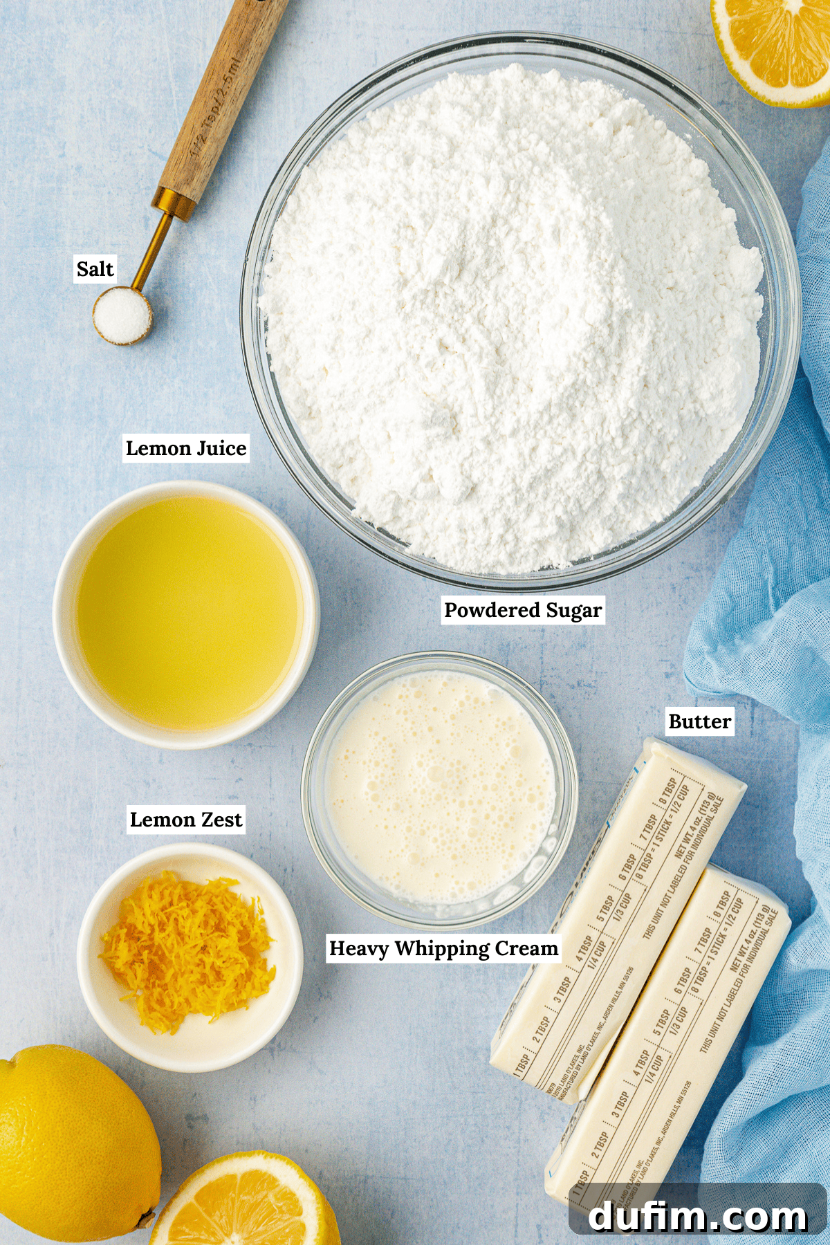 Overhead view of all the essential ingredients for making lemon frosting: unsalted butter, powdered sugar, fresh lemons for juice and zest, heavy whipping cream, and a pinch of salt.