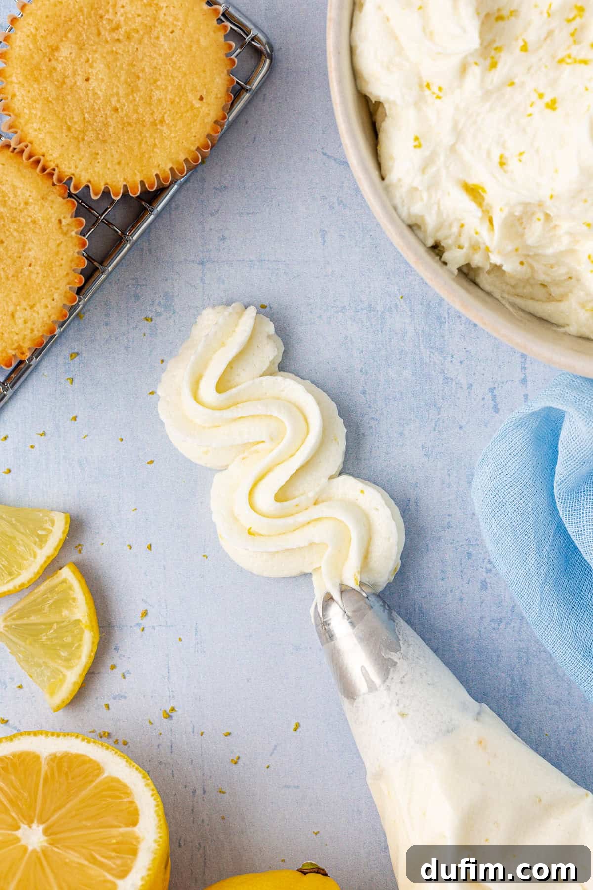 A piping bag filled with lemon frosting amidst a display of fresh lemon slices, cupcakes on a cooling rack, and a bowl of finished frosting.