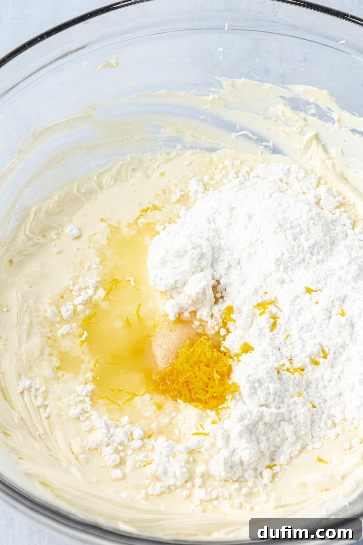 Ingredients for lemon frosting ready to be mixed in a clear glass bowl, showcasing powdered sugar, lemon juice, cream, and salt.
