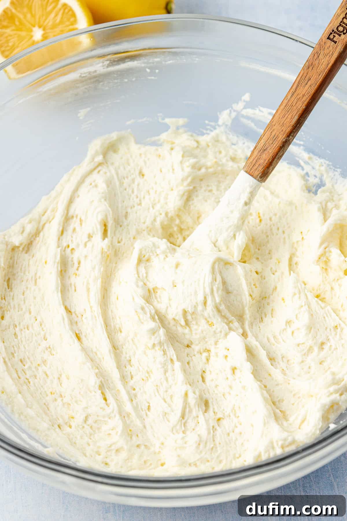A bowl of finished lemon frosting with a wooden spoon, and fresh lemons in the background, ready for use.