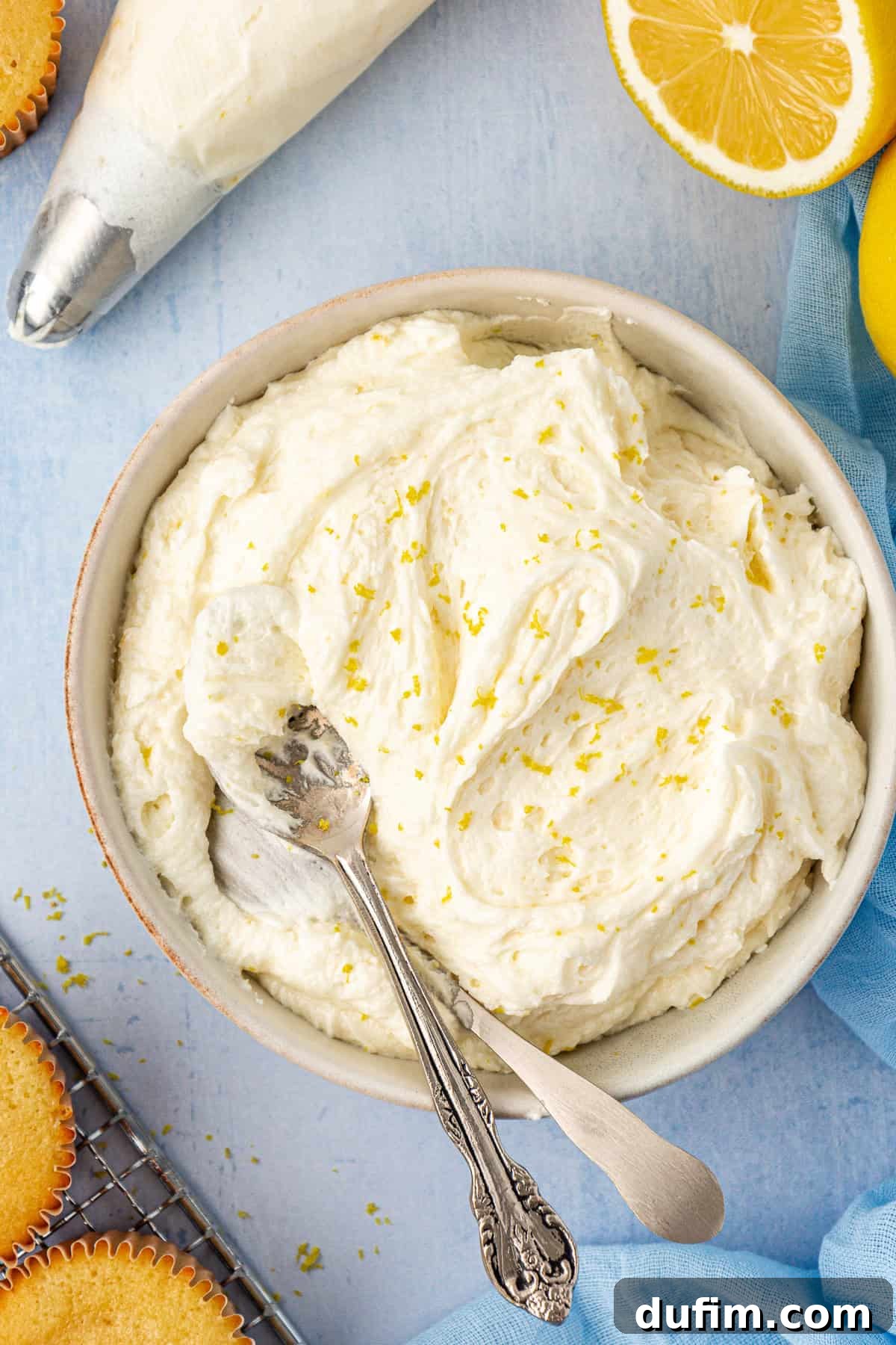 A beautifully arranged scene with a bowl of lemon frosting topped with zest, surrounded by a piping bag, frosted cupcakes, and fresh lemons.