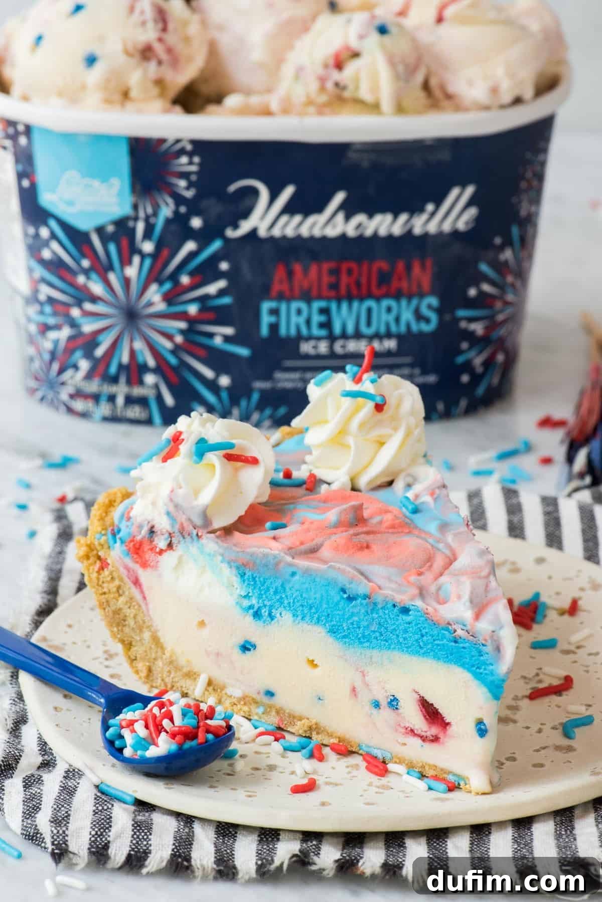 Slice of 4th of July red, white and blue ice cream pie on a white plate with a blue stripe napkin, with an American Fireworks ice cream container in the background.