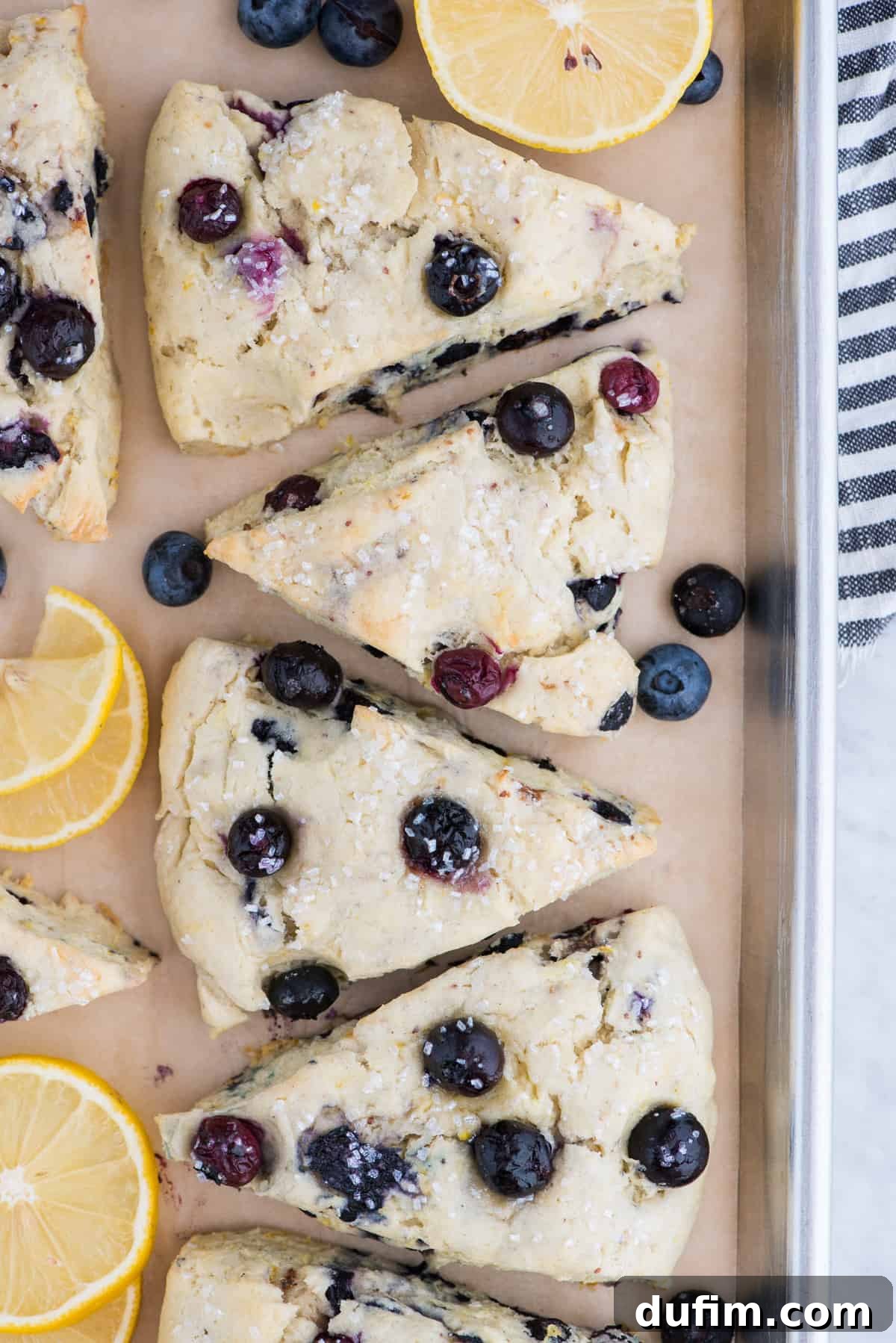 Perfectly baked lemon blueberry scones on brown parchment paper, accompanied by fresh lemon slices and ripe blueberries, highlighting their flaky texture.