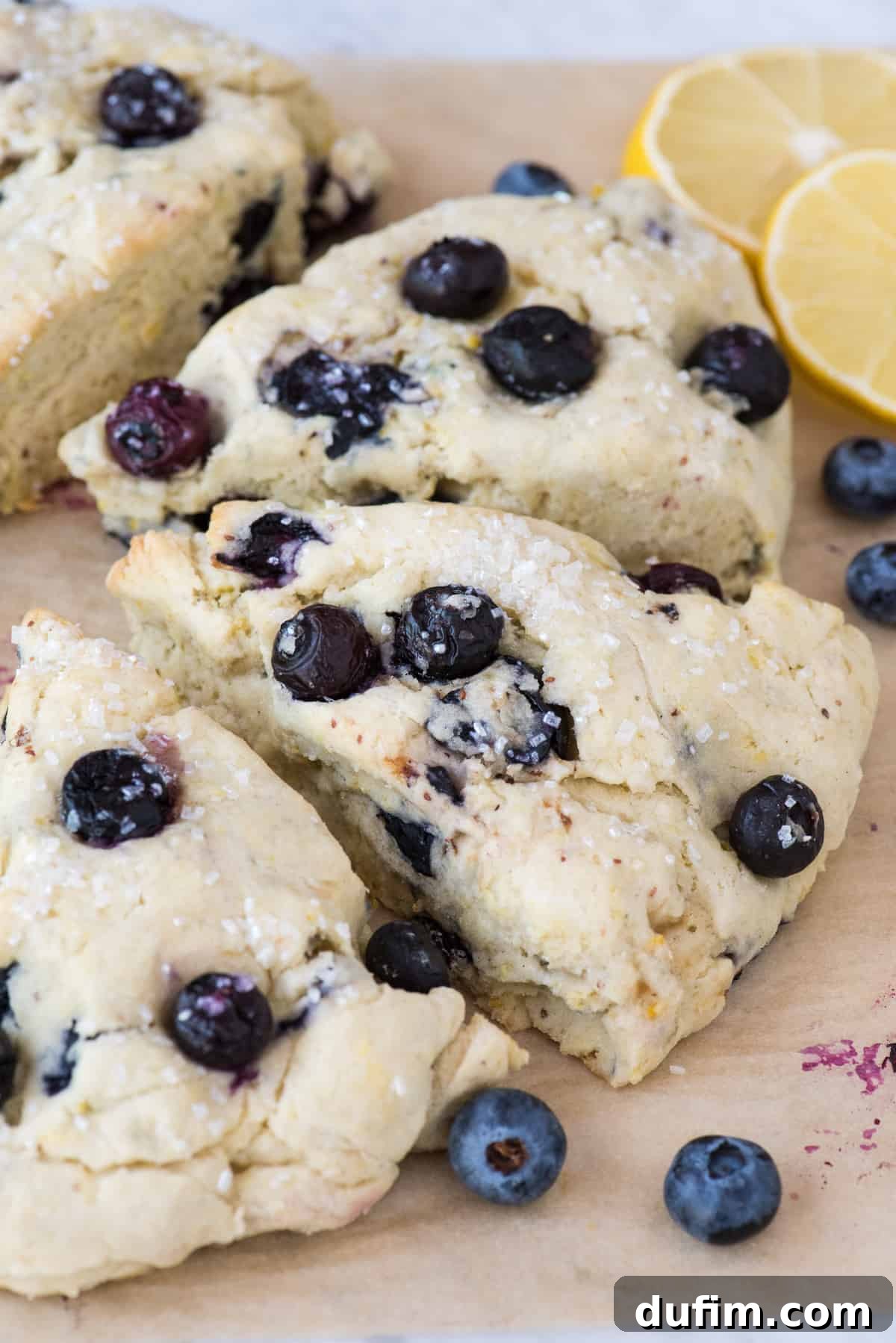 Close-up of baked lemon blueberry scones on brown parchment paper, artfully arranged with lemon slices and blueberries, highlighting their golden crust and tender interior.