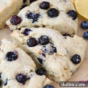 Three triangular lemon blueberry scones with a light dusting of sugar are arranged on parchment paper, surrounded by fresh blueberries and lemon slices in the background.