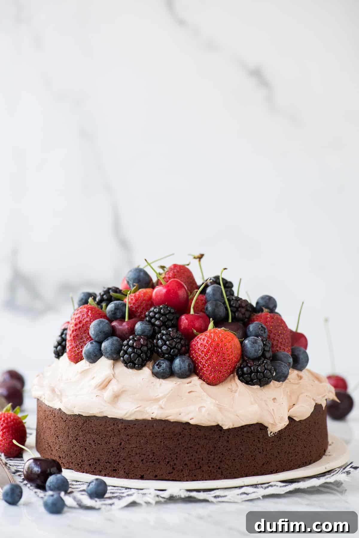 Velvet Chocolate Indulgence 2 Exquisite chocolate mud cake adorned with whipped frosting and a medley of fresh cherries, strawberries, and blackberries, elegantly presented on a white plate against a pristine white background.