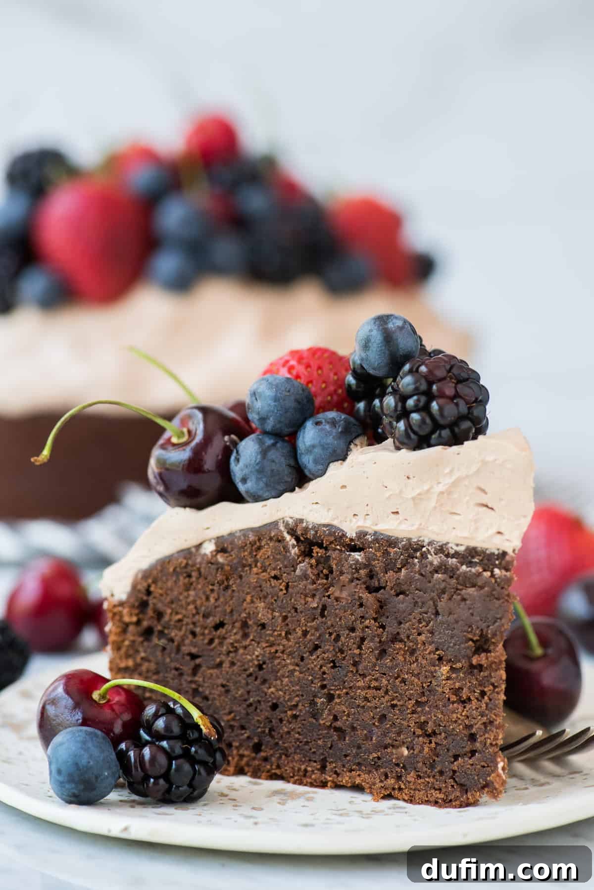 Velvet Chocolate Indulgence 4 A generous slice of chocolate mud cake, crowned with fluffy whipped frosting and garnished with fresh berries (cherries, strawberries, and blackberries), resting on a white plate against a clean white background, inviting a delicious indulgence.