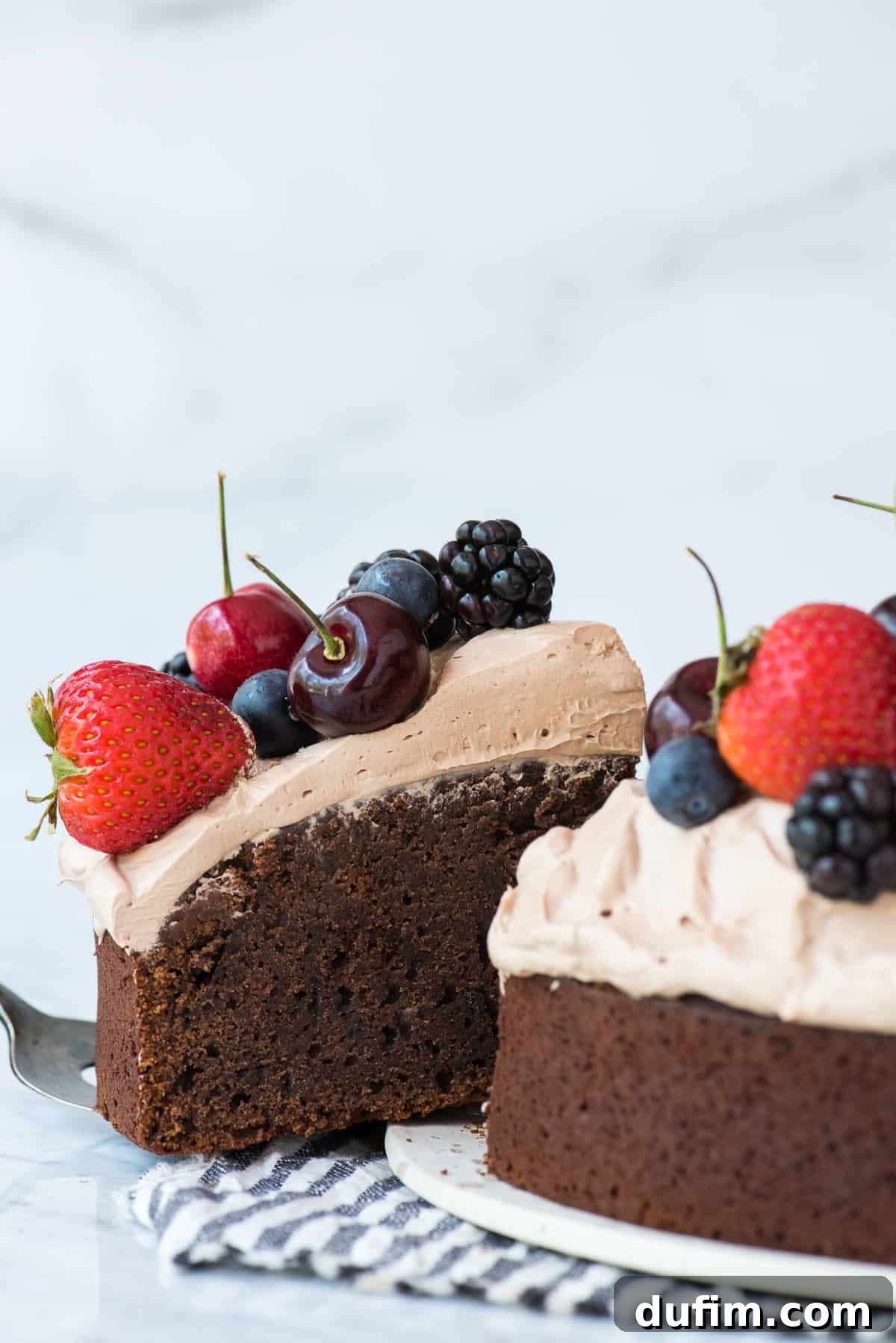 Velvet Chocolate Indulgence 6 A delightful slice of chocolate mud cake, generously topped with whipped frosting and fresh cherries, strawberries, and blackberries, is gently being removed from the main cake on a white background, highlighting its rich texture and inviting presentation.
