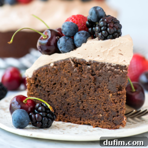 A slice of decadent chocolate mud cake with light brown frosting, topped with fresh blueberries, blackberries, cherries, and a strawberry, sits on a plate surrounded by extra berries.