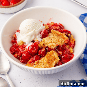A bowl of cherry cobbler crowned with a scoop of vanilla ice cream resembles a cherry dump cake. The dessert is vibrant red, punctuated by chunks of golden-brown crust. Nearby, a blue-checked napkin and two silver spoons rest on the pristine white surface.