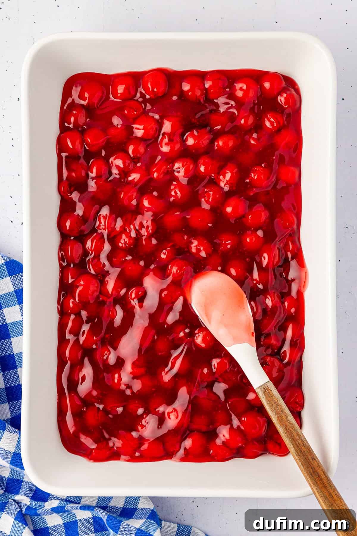 A white baking dish brims with glistening red cherry pie filling, hinting at a delicious cherry dump cake. A white spatula with a wooden handle rests on the edge of the dish, while a blue and white checkered cloth peeks out from the bottom left corner.