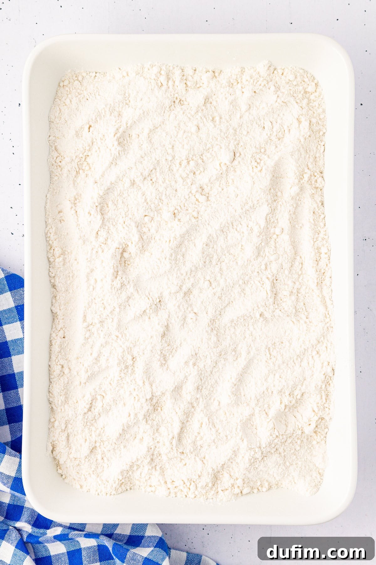 A rectangular white tray, perhaps poised for a cherry dump cake creation, is filled with a fine, white powdery substance—likely flour. It rests on a speckled surface, with the corner graced by a blue and white checkered cloth peeking in.