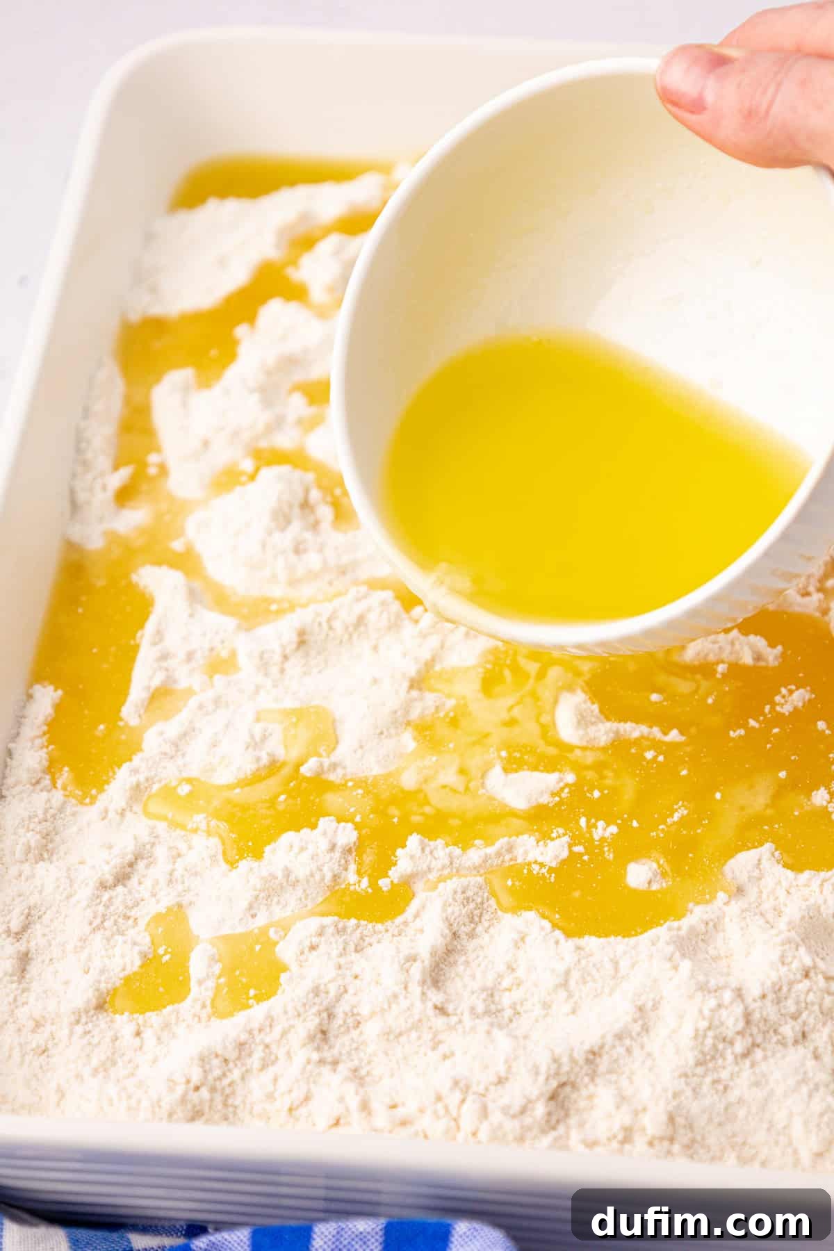 A hand pours melted butter from a white cup over a mix of flour in a rectangular white dish, setting the stage for a cherry dump cake delight. A blue and white striped cloth peeks out beneath the dish.