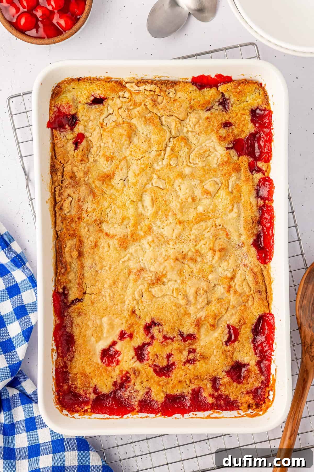 A rectangular baking dish holds a delectable cherry cobbler with a golden crust and red cherry filling peeking through the edges. Alongside, there’s a blue and white checkered cloth, a wooden spoon, and a bowl of cherries—reminiscent of the classic cherry dump cake.