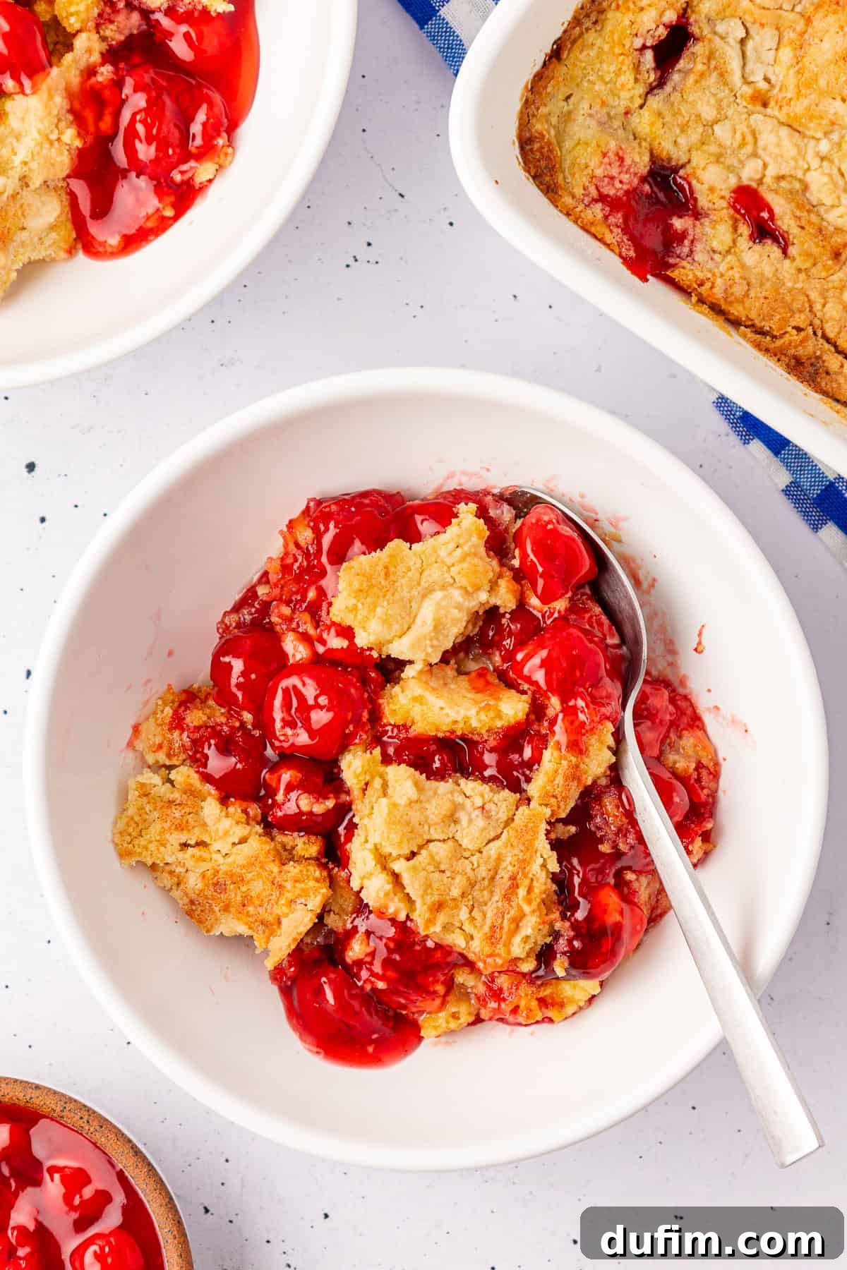 A bowl of cherry dump cake cobbler with a golden, crumbly crust is shown. A spoon rests in the bowl, surrounded by part of a baking dish and another bowl filled with tempting cobbler on a white speckled surface.