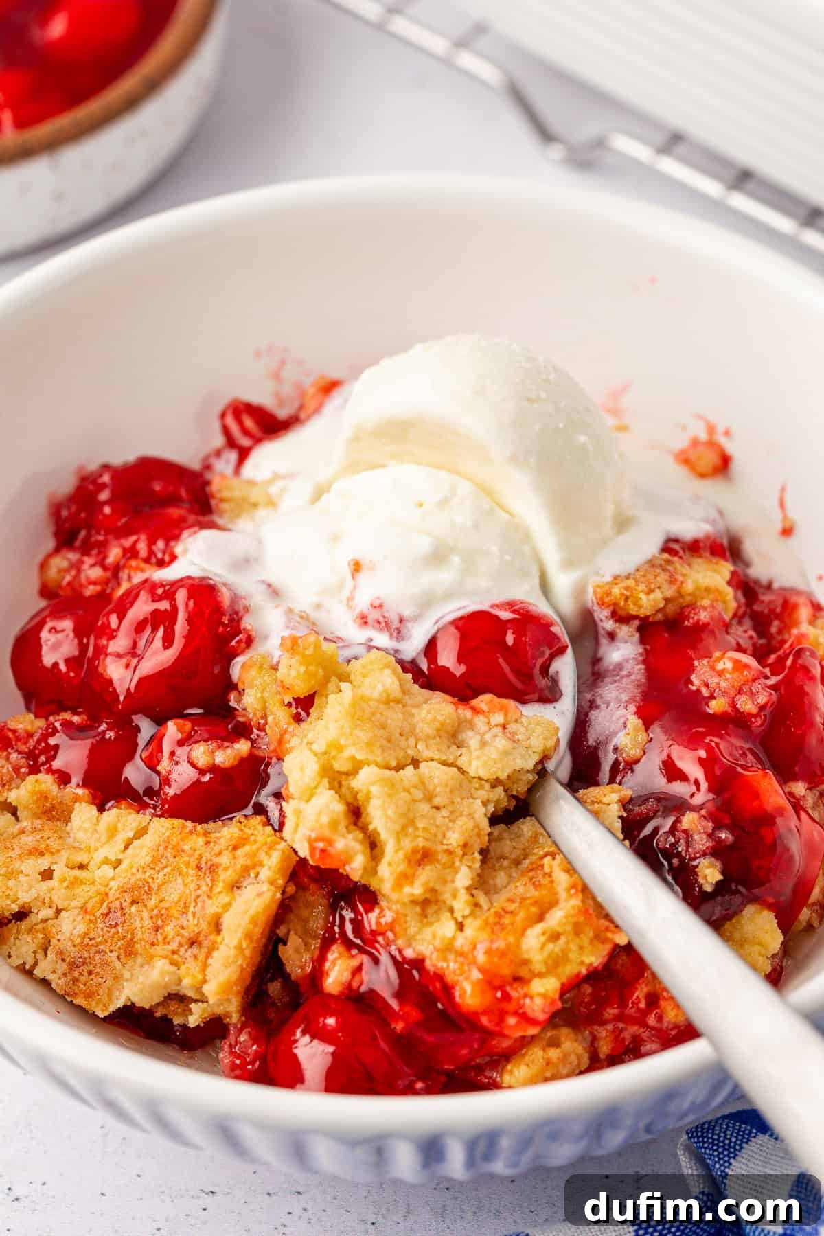 A close-up of a bowl filled with cherry cobbler and a scoop of vanilla ice cream on top. The cobbler, reminiscent of a cherry dump cake, boasts a golden-brown crust with vibrant red cherries, and a spoon rests in the dish. A hint of a blue cloth is visible at the bottom corner.