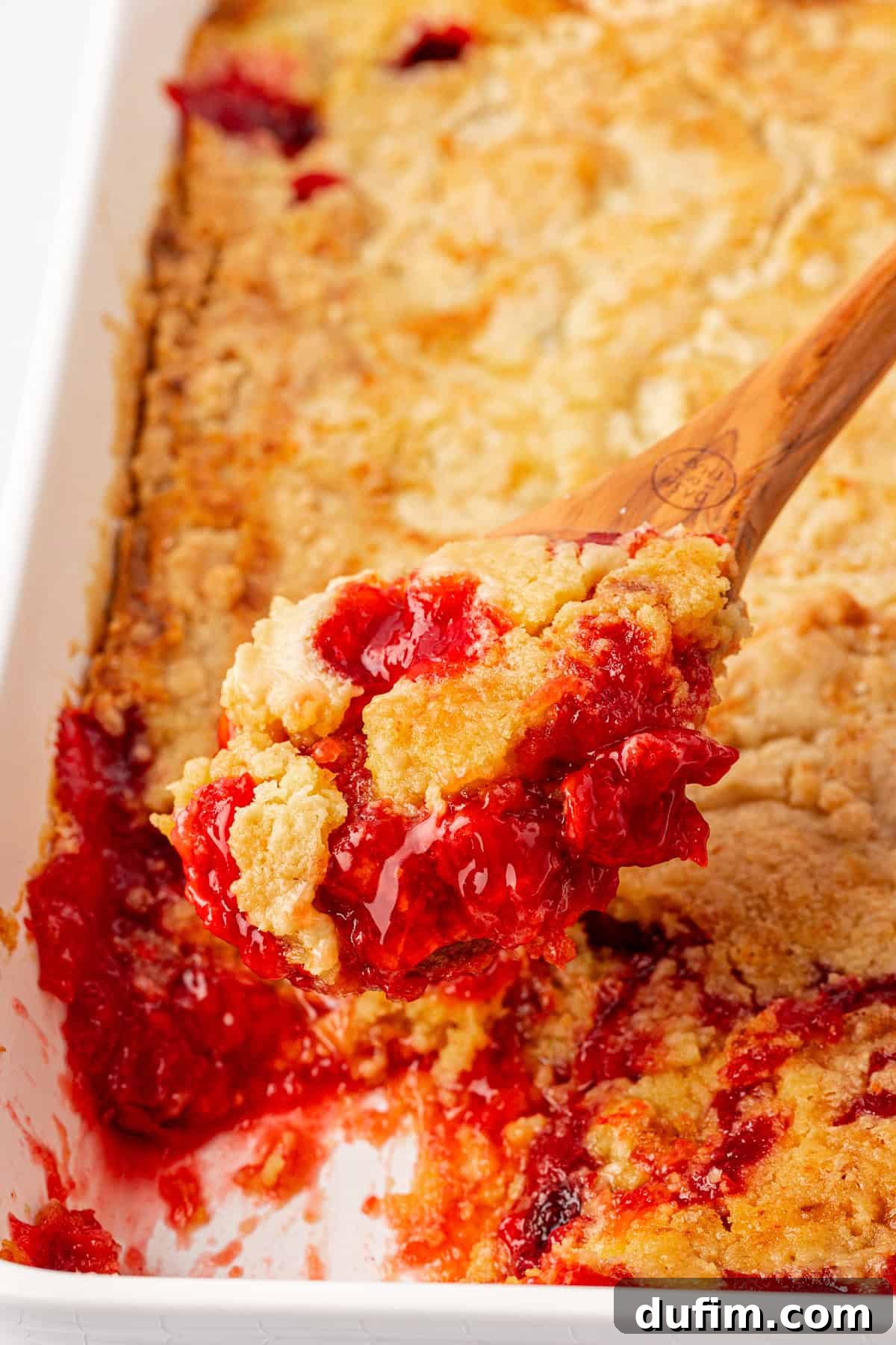 A close-up of a wooden spoon scooping a serving of cherry dump cake from a white baking dish. The dish showcases a golden-brown crust with vibrant red cherry filling bubbling below, giving it a warm and inviting appearance.