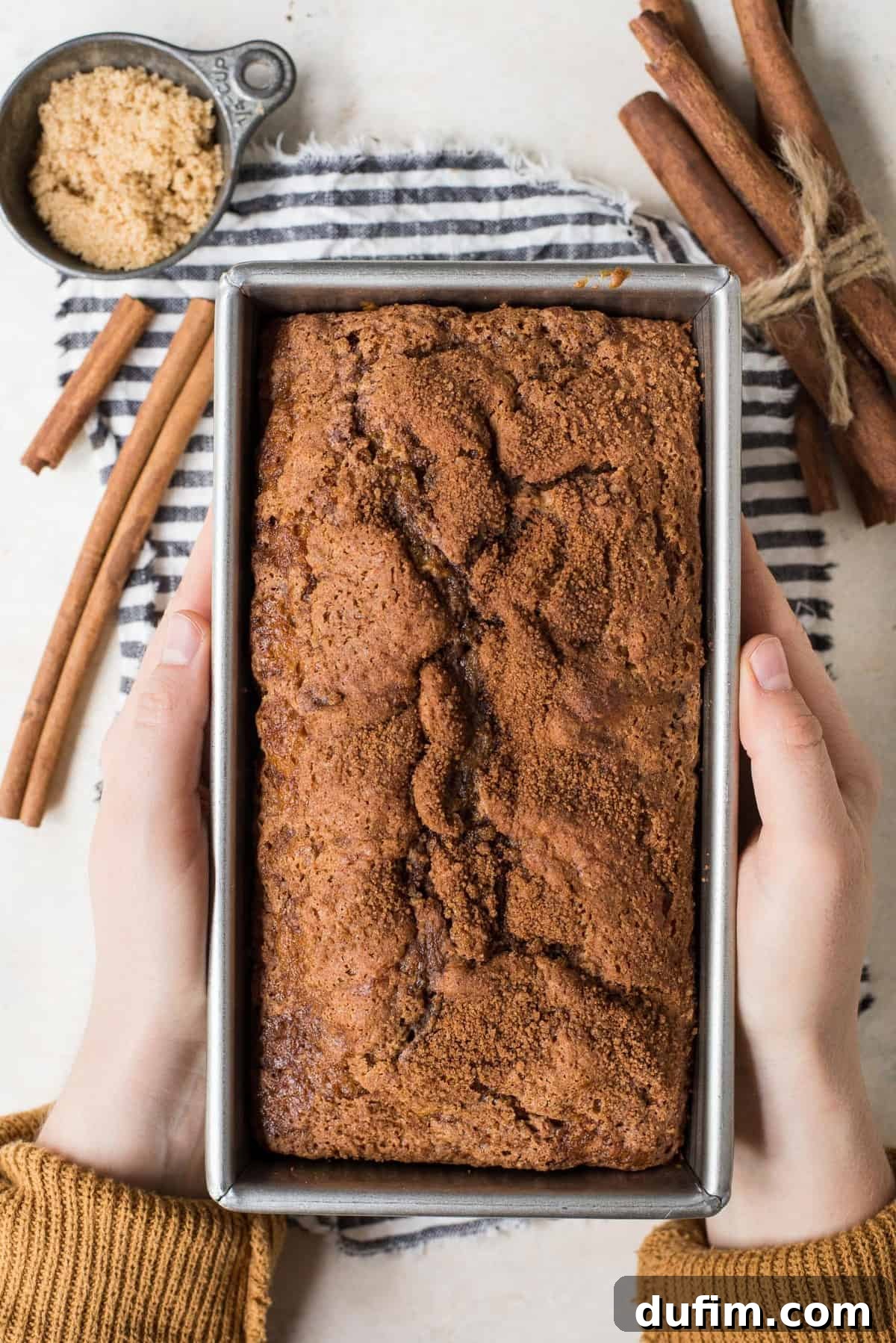 Hands gently holding a warm loaf of cinnamon swirl bread in a metal loaf pan, highlighting its golden-brown crust.