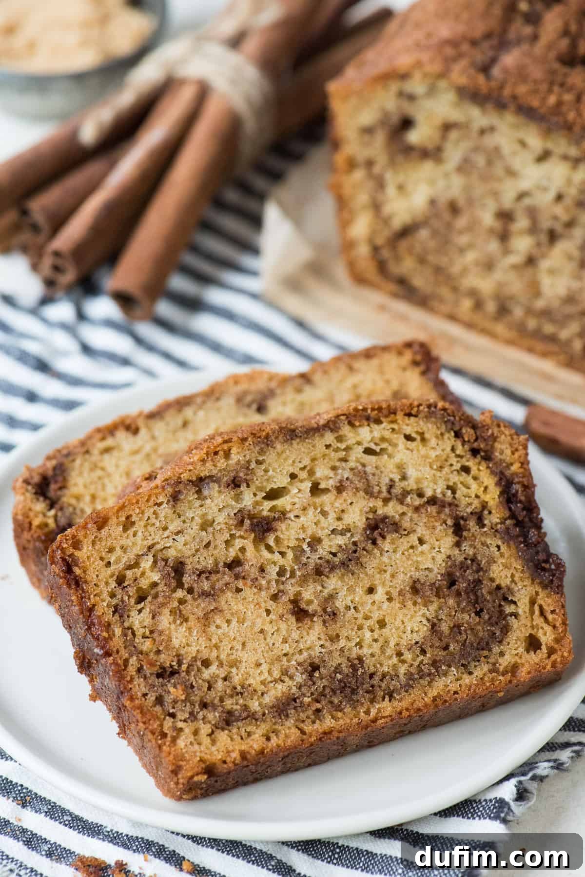 A cinnamon swirl bread with two slices already cut, revealing the intricate cinnamon pattern, presented on a blue striped linen.