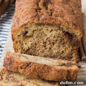 A loaf of cinnamon swirl bread with a golden-brown crust, partially sliced to reveal its marbled cinnamon pattern inside. Two cinnamon sticks and a striped cloth sit beside the inviting cinnamon swirl bread.