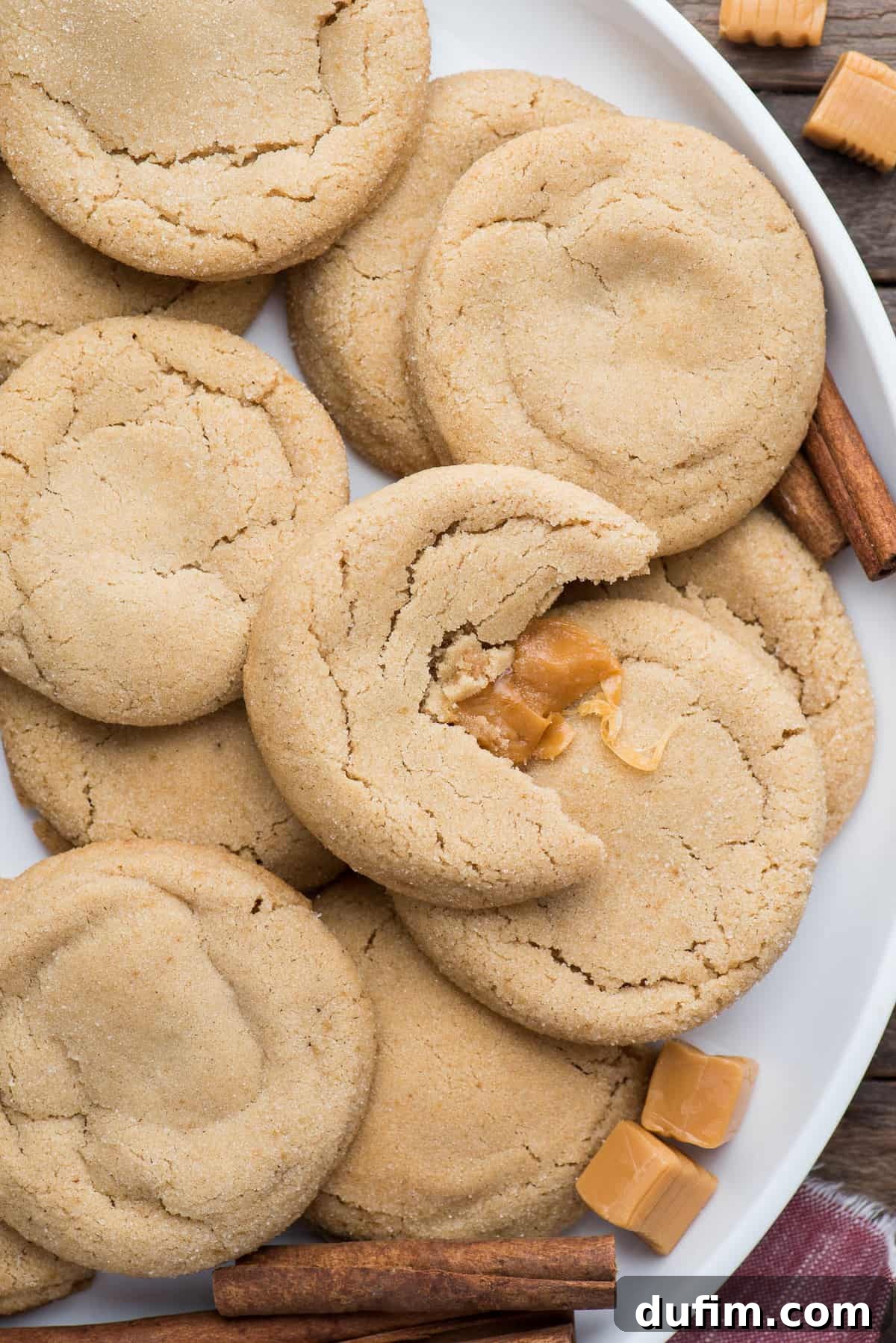 Soft and Chewy Caramel Apple Cookies with caramel centers on a white round platter, surrounded by loose caramel squares and cinnamon sticks.