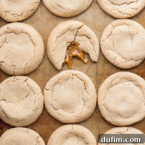 Twelve round caramel apple cookies on a baking sheet; one cookie in the center is broken, revealing gooey caramel inside.