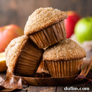 Three sugar-topped applesauce muffins are stacked on a wooden plate, with a few autumn leaves nearby and blurred apples in the background, creating a cozy fall atmosphere.