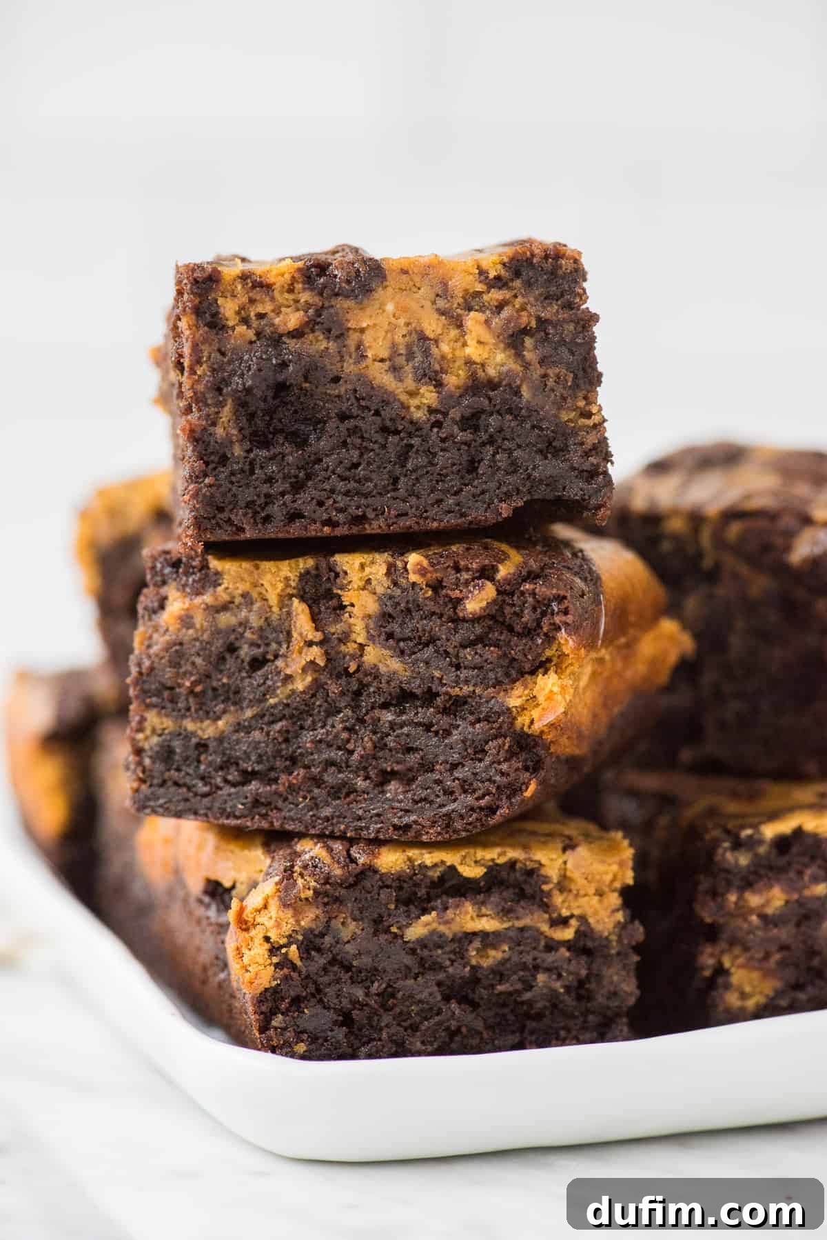 Pumpkin Cheesecake Swirl Brownies 3 Three decadent pumpkin cheesecake brownies stacked on a white tray, highlighting their distinct layers and rich texture.