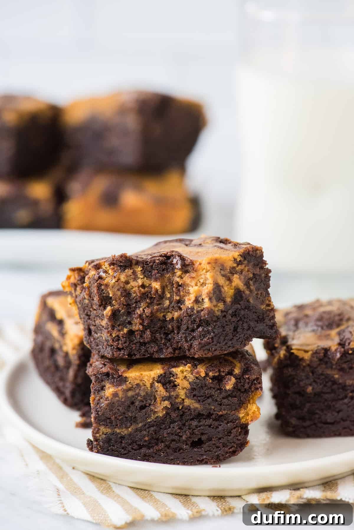 Pumpkin Cheesecake Swirl Brownies 6 Two pumpkin cheesecake brownies artfully stacked on a round cream plate with a striped linen underneath, ready to be served.