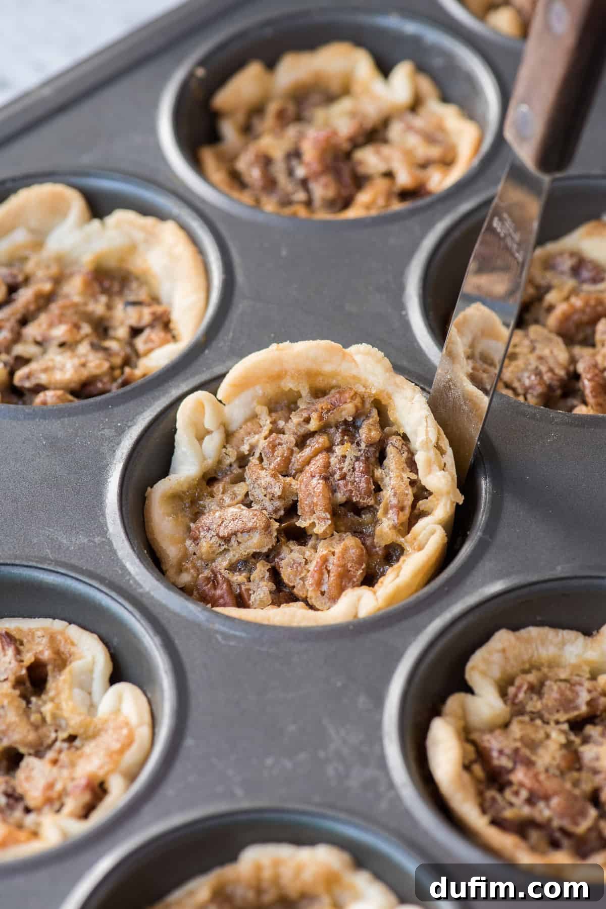 A perfectly baked mini pecan pie being carefully removed from a metal muffin pan with a metal spatula, highlighting its golden crust and inviting filling.