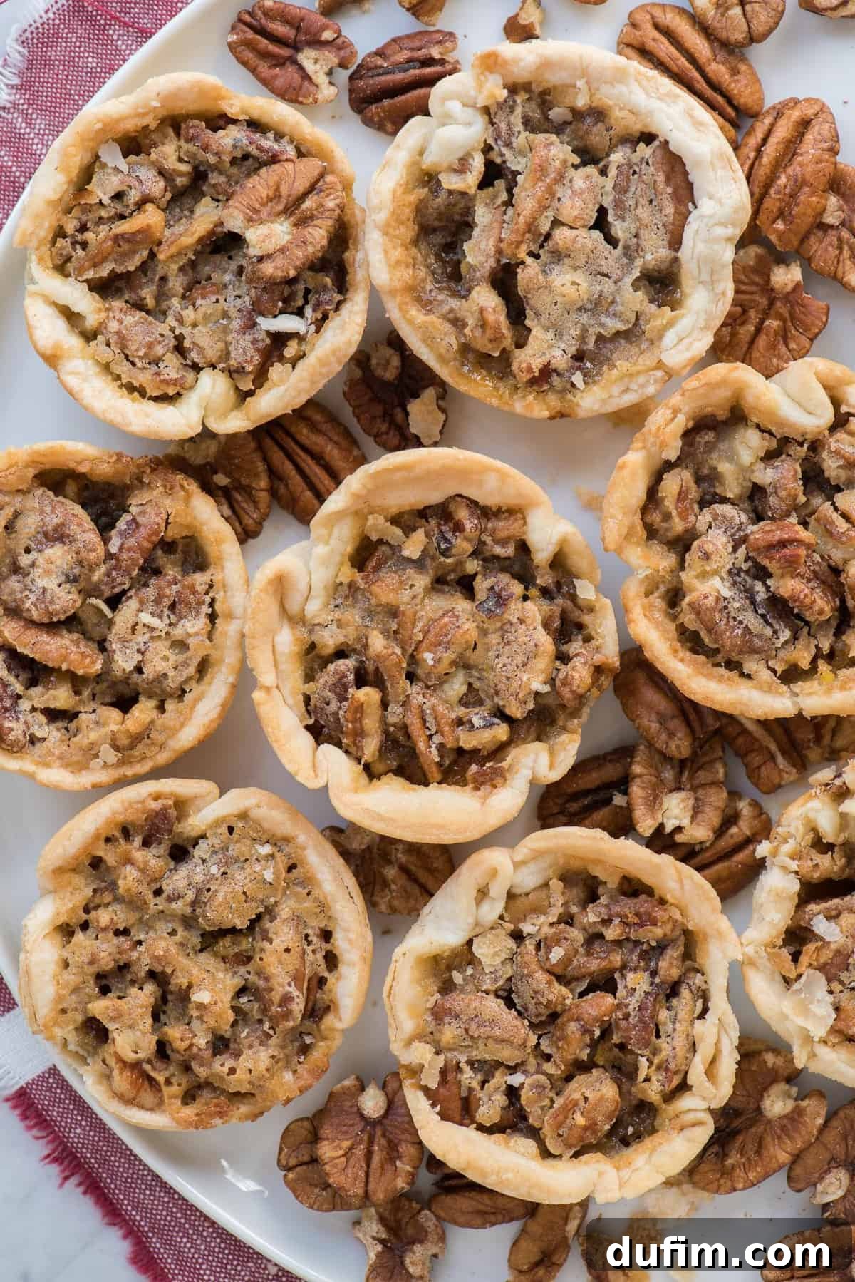 An aerial shot showcasing eight beautifully arranged mini pecan pies on a pristine white tray, accompanied by a festive red linen, perfect for a holiday spread.