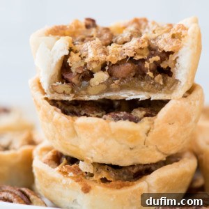 A close-up of three stacked mini pecan pies, with the top tart bitten to reveal the gooey, nut-filled filling inside the flaky pastry crust. A few more tarts and scattered pecans are visible in the background.