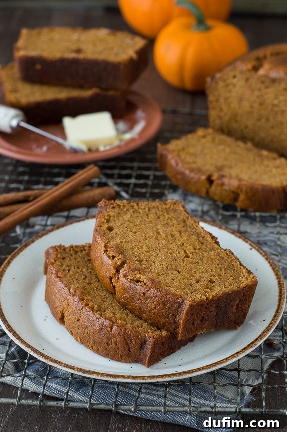 This Pumpkin Pound Cake recipe tastes just like Starbucks Pumpkin Pound Cake - takes 15 minutes to prep! Can be made in muffin, mini muffin or mini loaf pans. Two generous slices of moist Starbucks-style pumpkin bread, garnished with cinnamon sticks, resting on a small white serving plate atop a metal cooling rack. The background is softly blurred, evoking a cozy autumn atmosphere.