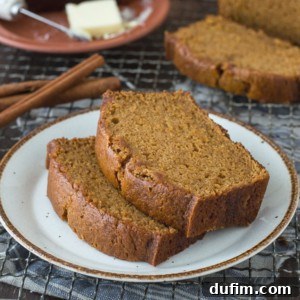 Two slices of moist, golden brown pumpkin bread—just like the Starbucks Pumpkin Bread Recipe—sit on a white plate, with cinnamon sticks nearby and a plate of butter in the background.
