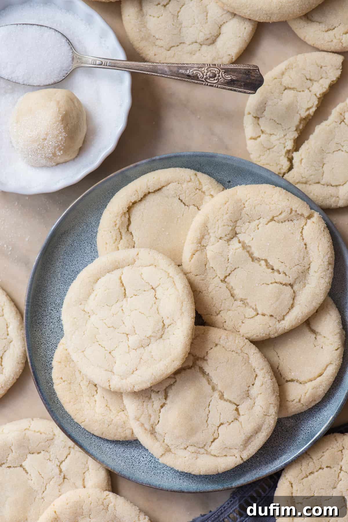 Freshly baked rolled sugar cookies piled on a blue plate, with a background of parchment paper and more cookies, showcasing their inviting texture.