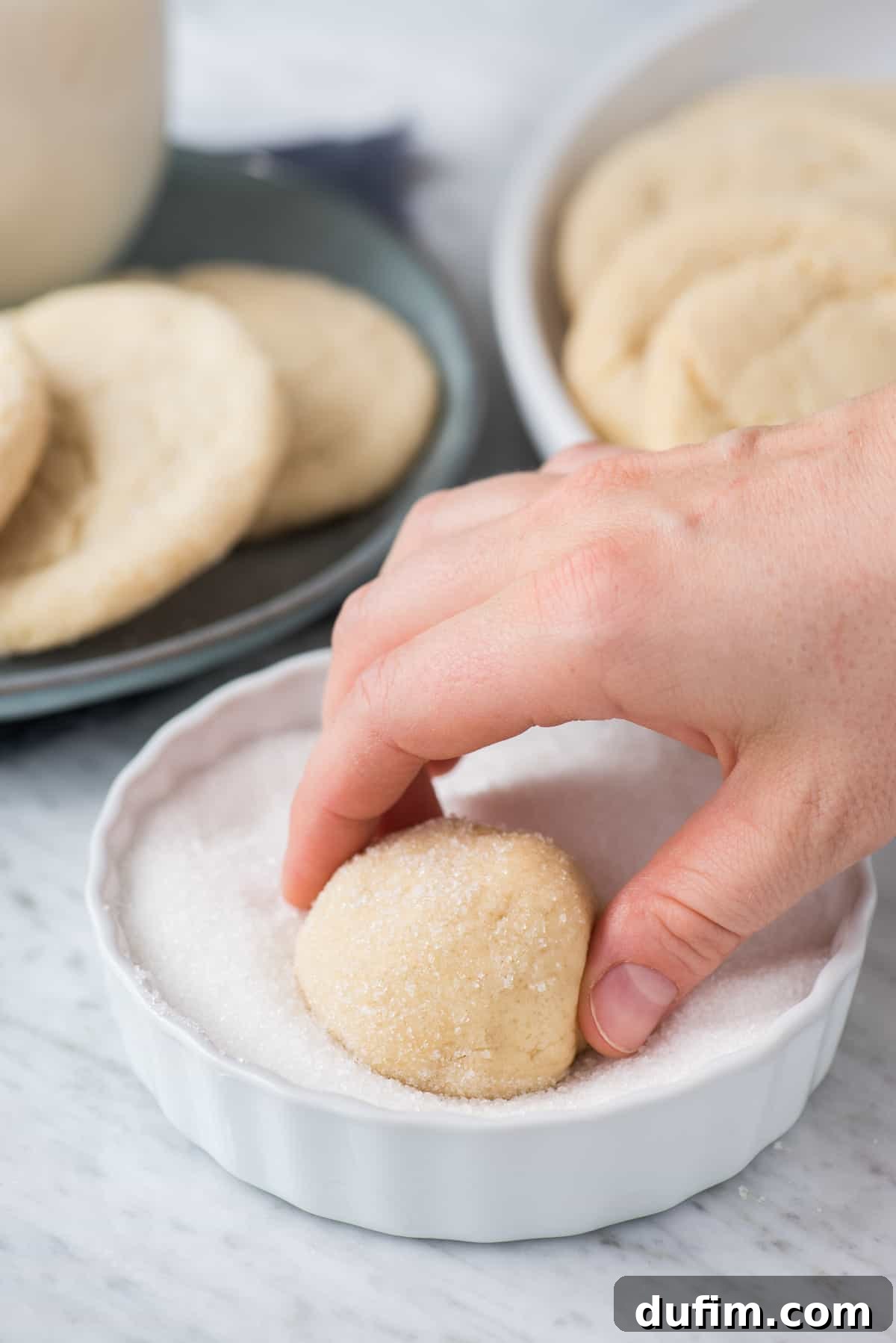 Close-up shot of a sugar cookie dough ball being rolled in granulated sugar in a bowl, showing the even coating process.