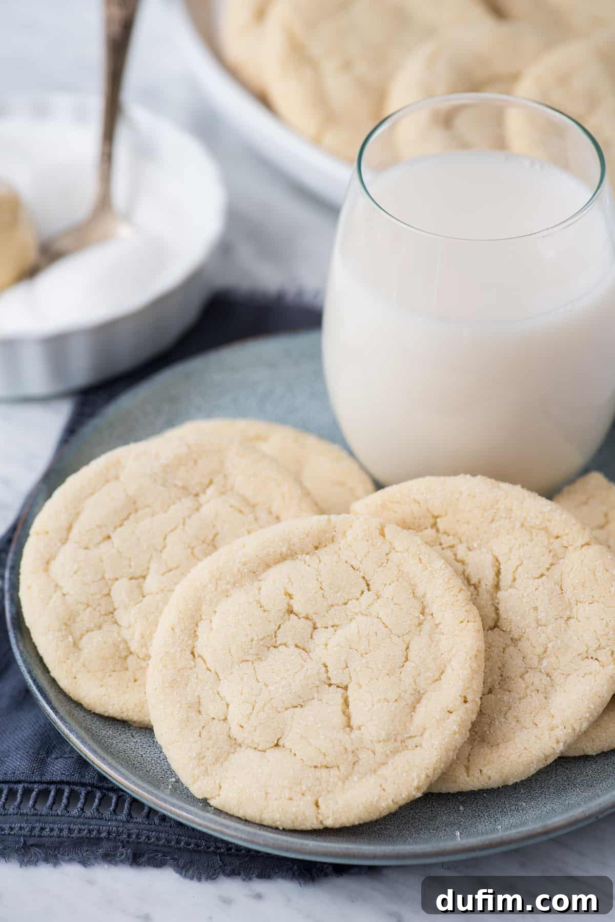 A blue plate holds a stack of rolled sugar cookies, with a glass of milk in the soft-focus background, suggesting a delightful snack.