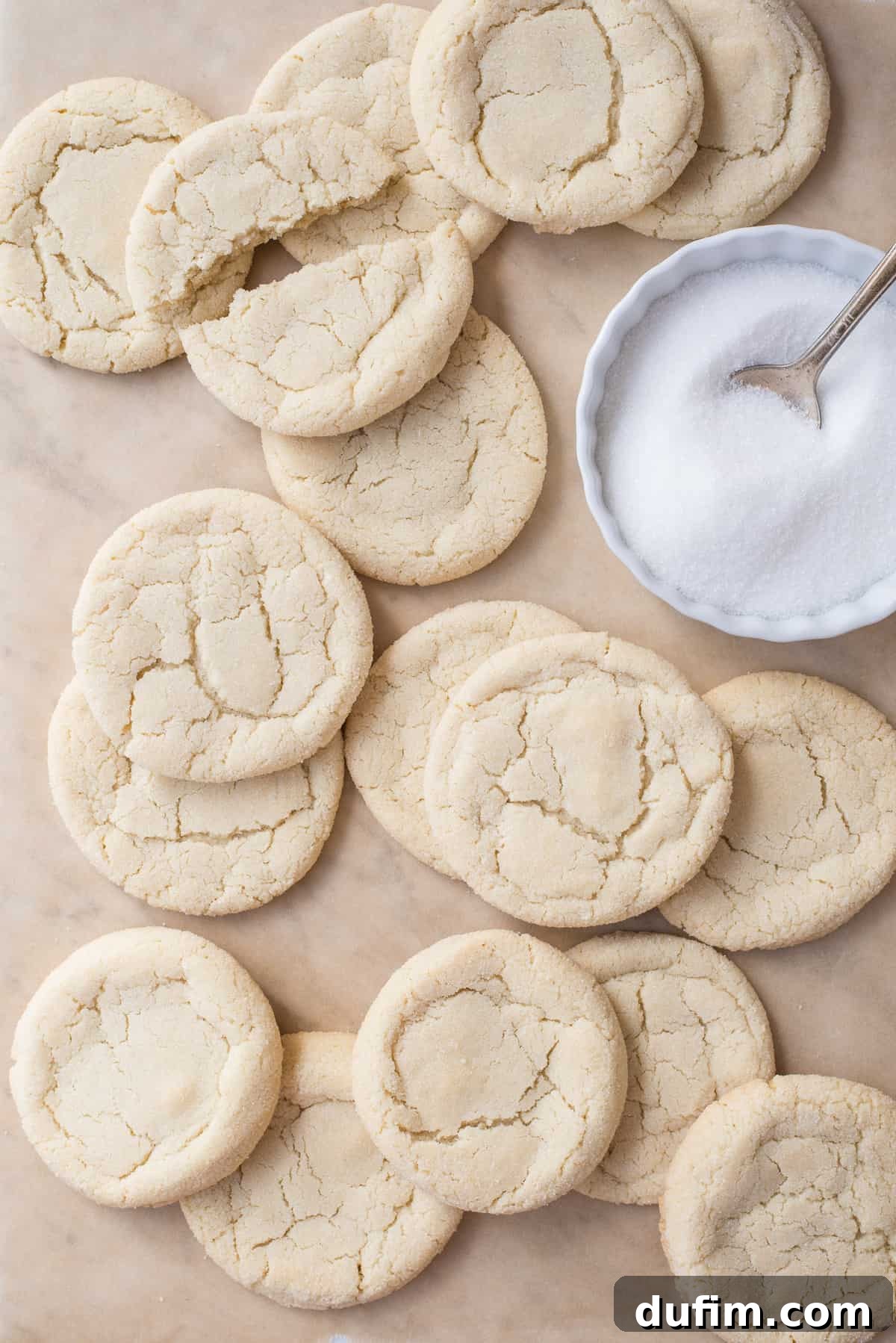 Scattered rolled sugar cookies on brown parchment paper with a bowl of sugar to the right, emphasizing their simple beauty.