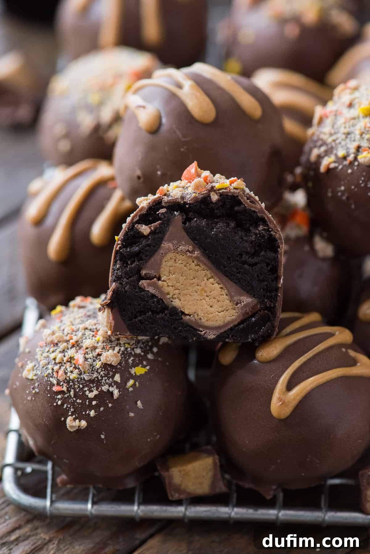 A top-down view of beautifully arranged Reese's Oreo Balls on a metal cooling rack, placed on a rustic wooden background. One ball in the foreground is cut open, showcasing the creamy peanut butter cup filling nestled inside the dark Oreo cookie exterior, inviting viewers to discover its delicious secret.