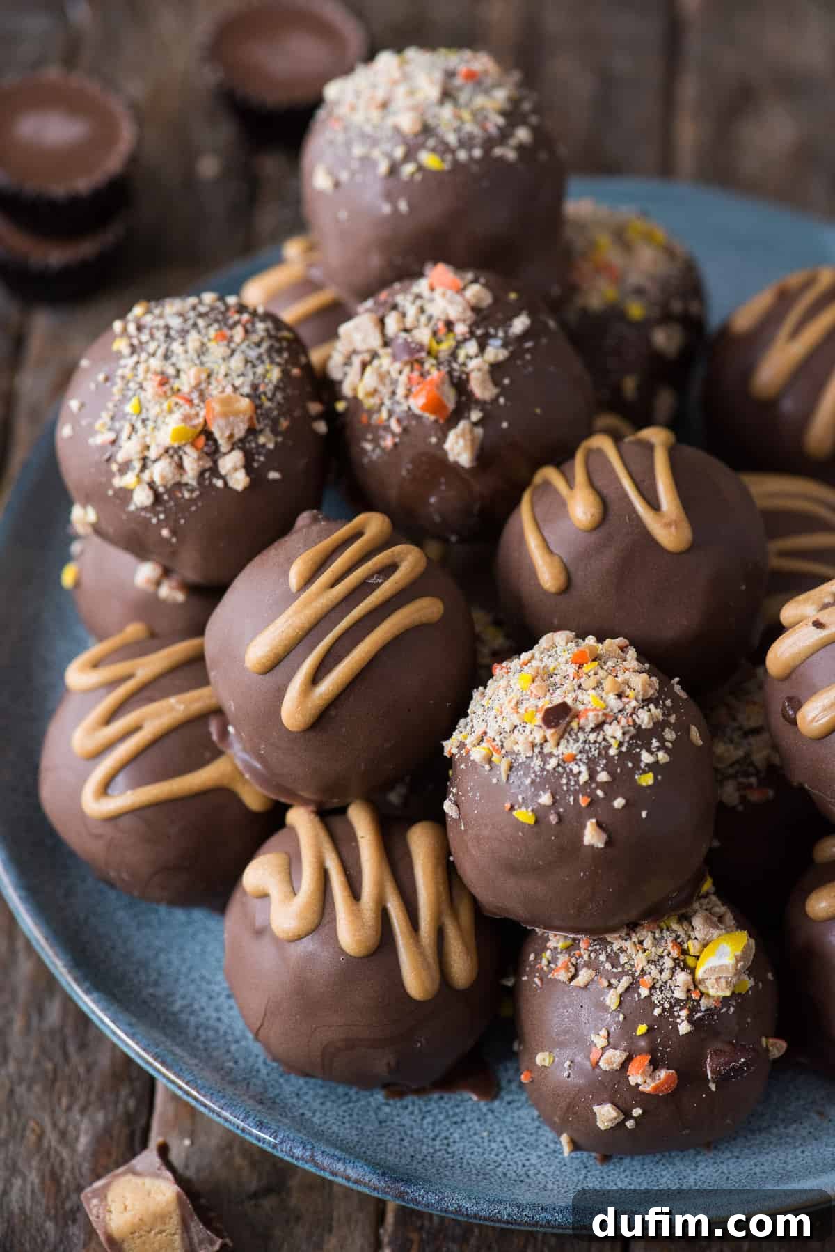 A vibrant blue plate holds a perfect display of chocolate-dipped Reese's Oreo Balls, each glistening with a smooth coating and topped with colorful Reese's Pieces and a delicate peanut butter drizzle, all set against a rustic wood background.
