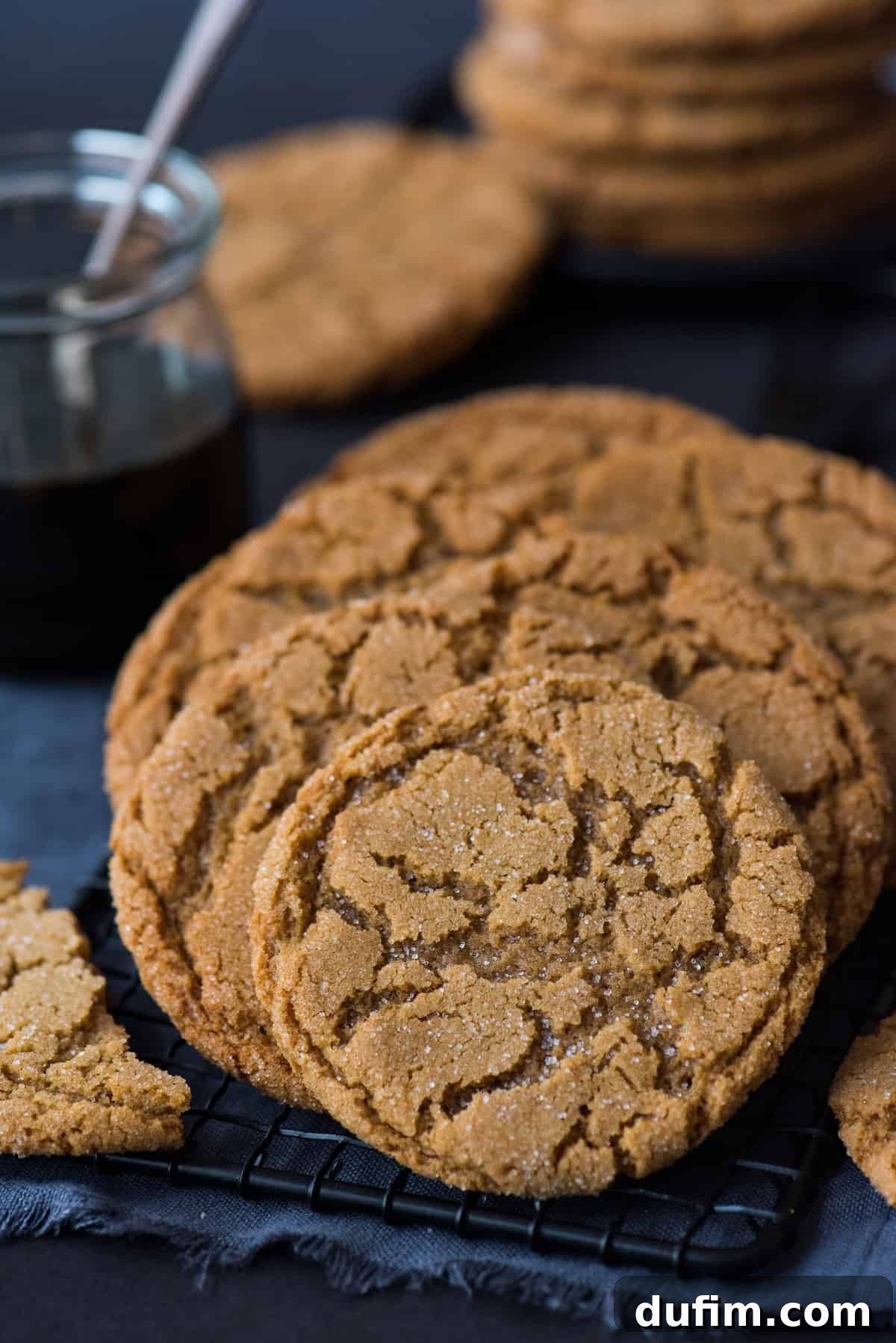 Ultimate Chewy Ginger Molasses Cookies 2 Molasses ginger cookies on a black wire rack, with additional cookies and a jar of molasses blurred in the background, creating a cozy and inviting baking scene.