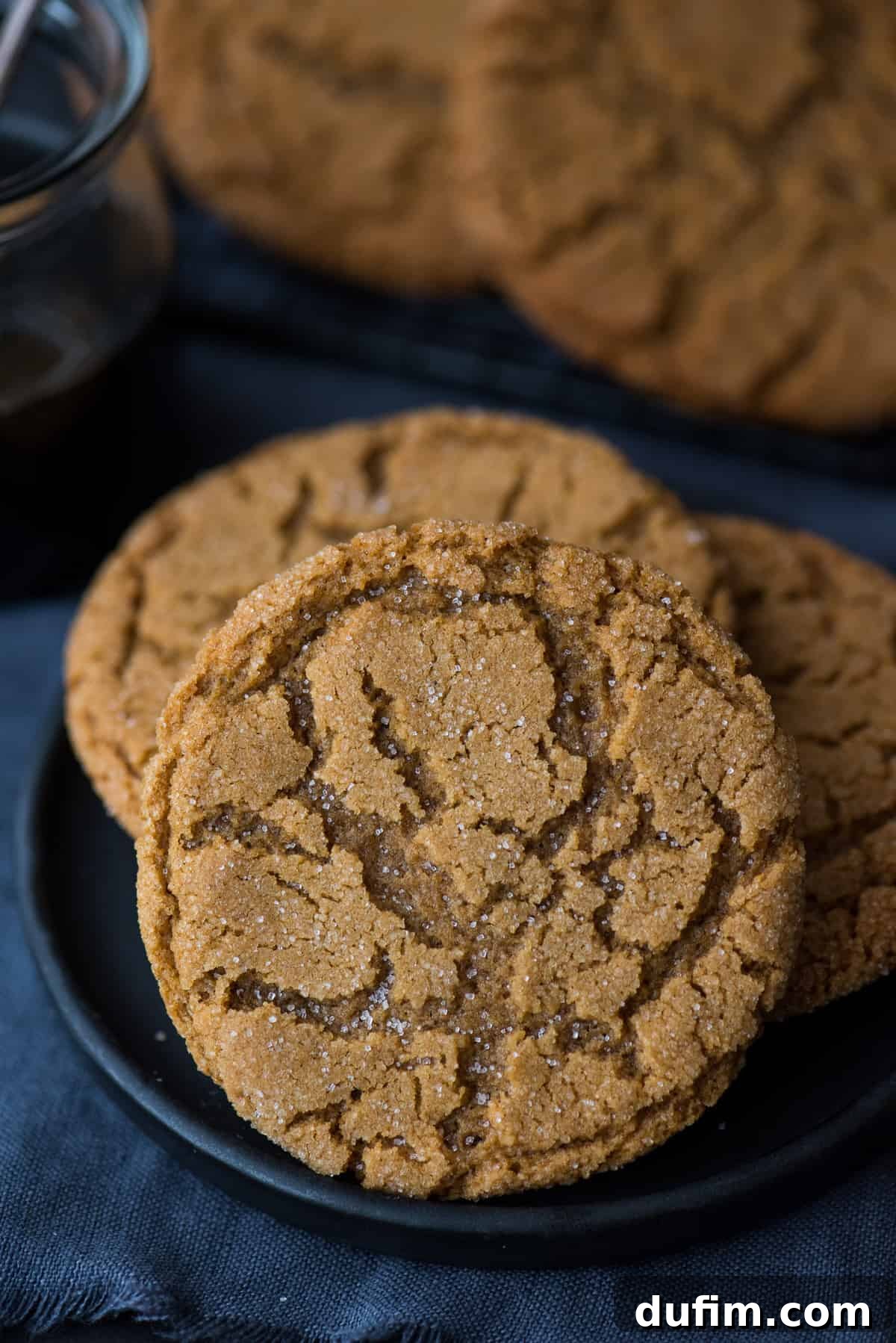 Ultimate Chewy Ginger Molasses Cookies 6 A single ginger molasses cookie on a dark plate, with a jar of molasses blurred in the background, highlighting the cookie's inviting appearance.