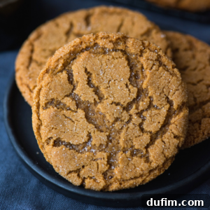 Close-up of a round, golden-brown ginger molasses cookie with a crackled surface and a sprinkling of sugar, resting on a dark plate with more cookies in the background.