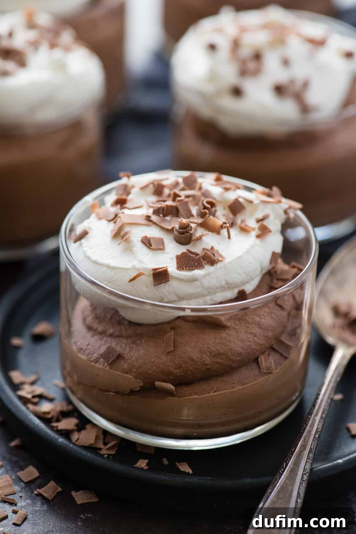 Silky chocolate mousse in a clear glass dessert bowl, elegantly topped with a swirl of fresh whipped cream and delicate chocolate shavings. The bowl rests on a dark plate, accompanied by a small metal spoon, highlighting its luxurious texture.